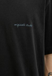 T-shirt en coton noir avec un texte bleu en cursive « originals studio » imprimé sur la partie gauche de la poitrine. Texture douce, design décontracté.