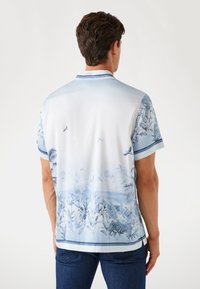 Korte mouwen shirt in lichtblauw met een tropisch ontwerp, featuring palmbomen en vogels. Gemaakt van gladde stof, met een kraag en trimaccenten.