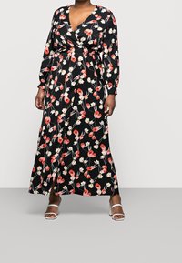 Robe maxi florale noire avec des motifs de fleurs rouges et blanches. Elle présente des manches longues, un décolleté en V et une taille ajustée. Portée avec des talons blancs.