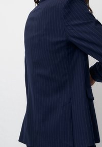 Femme portant un blazer bleu marine à fines rayures avec poche à rabat, debout devant un fond blanc uni, vue de côté.