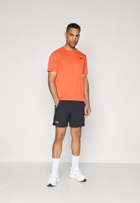 Camisa atlética de manga corta a rayas naranjas con un logo negro, combinada con pantalones cortos negros y zapatillas deportivas blancas. Diseño casual y suave.