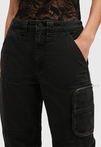 Zwarte cargobroek van denim met een gestructureerde afwerking. Bevat meerdere zakken, een ritszak aan de zijkant en een knoopsluiting bij de taille.