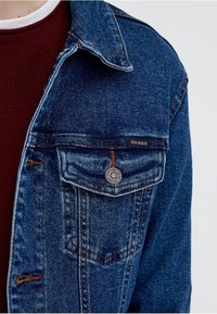 Jeansowa kurtka w ciemnoniebieskim kolorze z mocnym kołnierzem, z kieszenią na piersi z guzikiem oraz subtelnymi przetarciami na materiale.