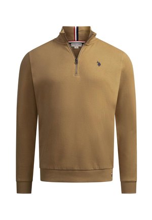Lächelnder Mann, der einen hellbraunen Pullover mit Viertelreißverschluss und kleinem schwarzem Logo auf der Brust sowie blaue Jeans trägt, Hand in der Tasche.