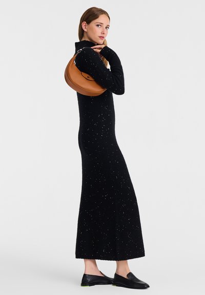 Schwarzes Strickkleid mit taillierter Silhouette und langen Ärmeln, ausgestattet mit silbernen Sprenkel-Details. Das Model hält eine braune Handtasche in geschwungener Form.