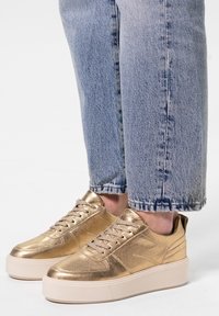 Gouden metalen sneakers met een gestructureerde afwerking, vetersluiting en een dikke witte zool. Draagt in combinatie met lichtblauwe, omgeslagen denim jeans.