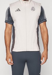 Chaleco ligero tipo zip, con frente gris claro y paneles laterales gris oscuro, textura acolchada, con los logos de Adidas y Real Madrid.