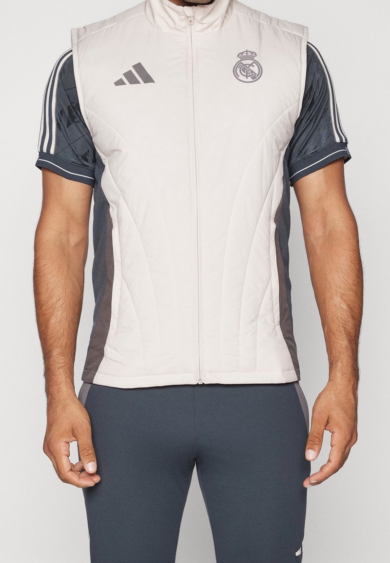 Chaleco ligero tipo zip, con frente gris claro y paneles laterales gris oscuro, textura acolchada, con los logos de Adidas y Real Madrid.