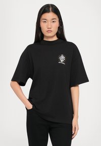 T-shirt in cotone nero con maniche corte, scollo rotondo e una grafica frontale che presenta un design di cuore e corona con accenti bianchi e dorati.