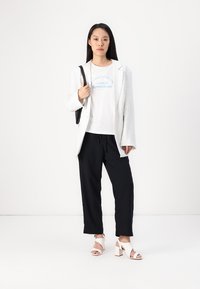Blazer bianco strutturato su una t-shirt bianca con testo blu, pantaloni neri larghi e sandali bianchi con tacco grosso, mentre tiene una borsa nera.