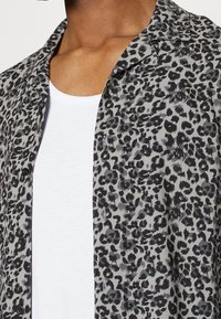 Košile s leopardím potiskem v černé a šedé barvě, vyrobená z lehkého materiálu, má límeček a knoflíkovou přední stranu, nošena přes bílou košili.