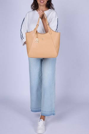 Vrouw draagt een wit topje, lichtblauwe spijkerbroek en witte sneakers, en houdt een grote beige lederen handtas vast voor een lichte achtergrond.