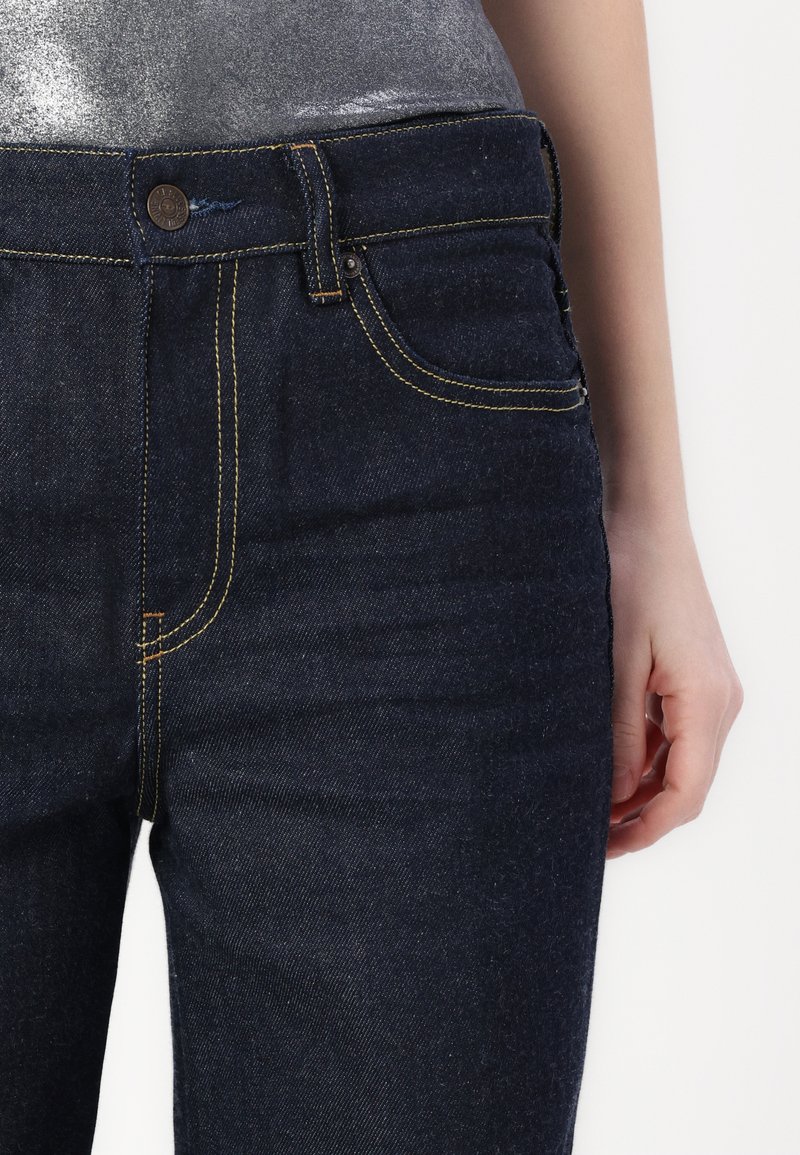 Dunkelblaue Denim-Jeans mit gelben Nähten, einem Knopfverschluss vorne und einer Seitentasche. Die Textur wirkt glatt mit einem leichten Glanz.