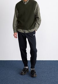 Jersey verde oliva sobre una camisa de botones a rayas, combinada con pantalones de chándal negros y zapatos negros de cordones con suela gruesa.