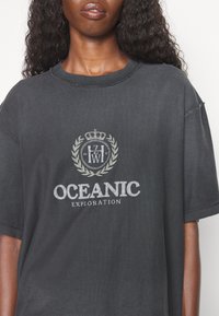 Černé oversized tričko s texturovaným grafickým prvkem a detaily vavřínu a koruny. Text zní "OCEANIC EXPLORATION" s opotřebeným vzhledem.
