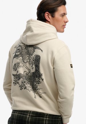 Hombre con sudadera con capucha beige con grullas negras de estilo japonés y diseño floral en la parte trasera, de perfil con las manos en los bolsillos.