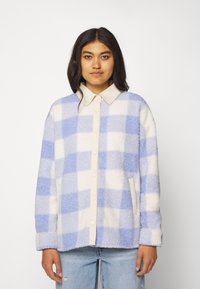 Vrouw in een blauw-wit geruit fleece jack met een beige kraag en knopenlijst, gecombineerd met lichtblauwe ripped jeans.