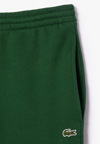 Joggers en coton vert avec une taille élastique, des poches latérales et un petit logo brodé en bas à droite. Texture lisse.