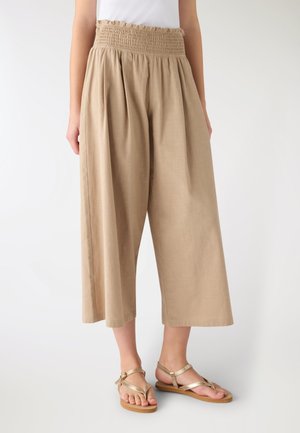 Donna che indossa pantaloni beige a gamba larga e taglio corto con vita arricciata, top bianco e sandali bassi dorati con cinturini, in piedi su uno sfondo chiaro.