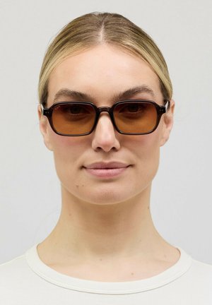 Woman with blonde hair wearing dark brown rectangular sunglasses and a white crewneck top, neutral background.
