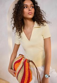 Hellgelbes, kurzärmliges Polo mit V-Ausschnitt. Sie hält eine mehrfarbige, gestrickte Tasche mit orangefarbenen, roten und natürlichen Tönen, die braune Träger hat.