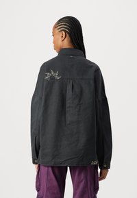 Giacca nera oversize con colletto strutturato, piega centrale sul retro e dettagli ricamati a sole. Presenta maniche ampie e chiusura con bottoni a pressione.