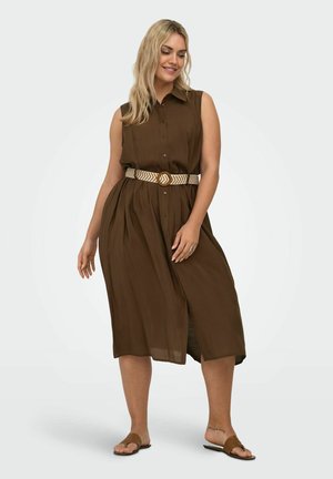 Femme portant une robe midi marron sans manches boutonnée avec une ceinture à motifs et des sandales plates marron, debout devant un fond uni.