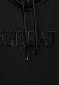 Svart huvtröja i mjukt tyg med stor svart broderad text "GREAT" och en mindre skriven text nedan. Inkluderar justerbara dragsko med metalldelar.