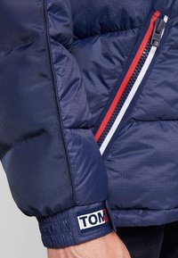 Granatowa kurtka puchowa o pikowanej fakturze. Posiada czerwono-biało-niebieski zamek błyskawiczny, elastyczne mankiety oraz naszywkę z logo na rękawie.