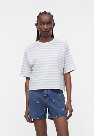 Νεαρή γυναίκα με πλεκτά μαλλιά που φοράει ένα γαλάζιο και λευκό ριγέ μπλουζάκι και τζιν σορτς με μικρή λευκή κεντητή διακόσμηση με λουλούδια.