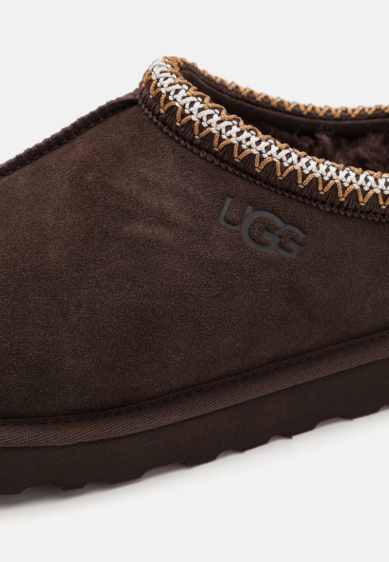 Bruine suède pantoffel met een getextureerd oppervlak, contrasterende gevlochten accent aan de kraag en UGG-logo op de zijkant geperst. Rondvormig.