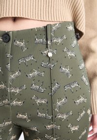 Groene broek met een herhaald zebrapatroon in zwart en crème, met een knoopsluiting aan de voorkant en een gladde, lichtglanzende textuur.