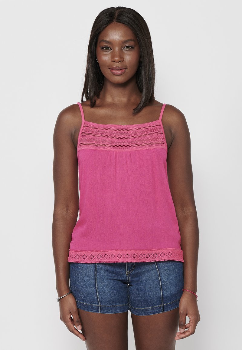 Débardeur rose avec des détails en dentelle, en tissu léger et avec des bretelles ajustables. L'ourlet présente un motif œillet décoratif. Porté avec un short en jean.