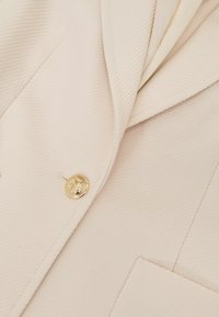 Beežist pintsak tekstuurse kangaga, millel on kuldne nööp ja sälguga krae. Sisaldab rinnataskut ja peeneid diagonaalseid triibusid.