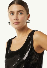 Czarny top bez rękawów z cekinowym przodem, okrągłym dekoltem i szerokimi ramiączkami. Srebrne kolczyki w kształcie kół dopełniają strój, dodając mu blasku.