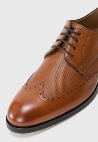 Zapato de vestir de cuero marrón con diseño perforado en la parte superior, punta afilada y cordones oscuros en contraste. Presenta detalles decorativos en brogue.