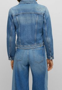 Žena oblečená v denimové bundě středně modré barvy a dvoubarevných širokých kalhotách, ze zadu, na jednobarevném šedém pozadí.