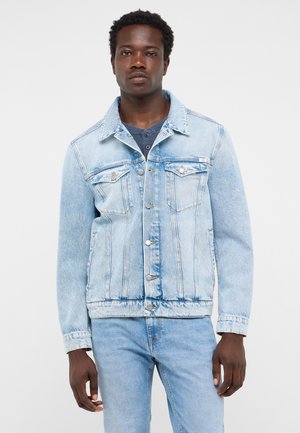 Mann trägt eine hellblaue Jeansjacke und Jeans, steht vor einem schlichten Hintergrund und schaut mit neutralem Gesichtsausdruck nach vorne.