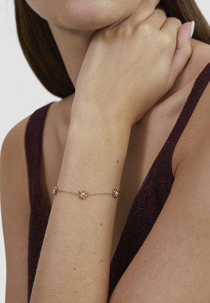 Delicate gold bracelet with small pearl clusters worn on wrist of person in sleeveless dark purple textured top.