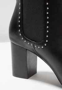Černá kožená kotníková bota s masivním čtvercovým podpatkem, boční elastickou vsadkou a kovovými nýty podél švu pro dekoraci.