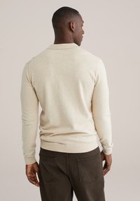 Mann trägt beige Langarmpullover und dunkelbraune Hose, steht mit dem Rücken zur Kamera vor einem einfarbigen Hintergrund.