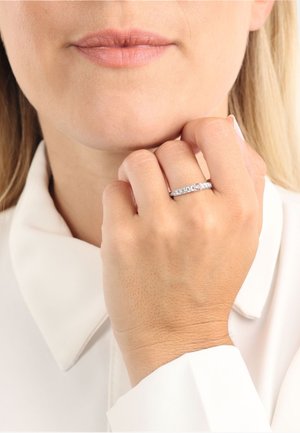 Anello in oro bianco con piccole pietre preziose trasparenti disposte in fila, indossato su una mano di carnagione chiara, su uno sfondo di morbido colletto bianco.