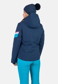 Dunkelblaue wasserdichte Jacke mit Kapuze, mit pinken und türkisfarbenen Streifen an den Ärmeln; kombiniert mit türkisfarbenen Hosen.