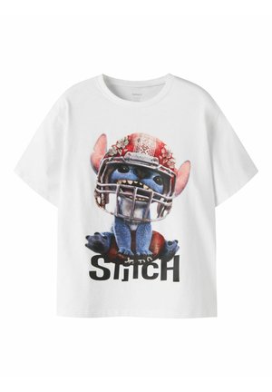 T-shirt blanc avec un graphisme du personnage bleu Stitch portant un casque de football rouge et assis sur un ballon de football marron, avec le texte "STITCH" en dessous.