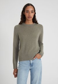 Jeune femme aux cheveux bouclés portant un pull en tricot vert olive et un jean bleu clair, une main dans la poche, fond neutre.