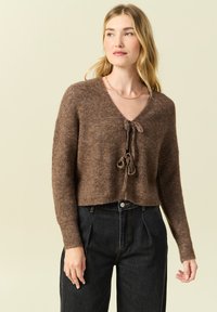 Maglione lavorato a maglia marrone con scollo a V e chiusura frontale a fiocco. Ha maniche lunghe e un design cropped, abbinato a jeans neri a vita alta.