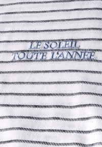 Bílá látka s tenkými námořnickými pruhy. Obsahuje vyšívaný text "LE SOLEIL TOUTE L’ANNEE" v jasně modré barvě. Měkká textura s viditelným stehem.