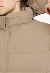 Beige puffjacka med hög krage, dragkedja och subtil logodetalj. Ytan har en slät, matt textur med horisontell sömnad.
