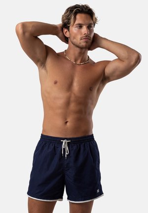 Mężczyzna bez koszulki, z jasnym zarostem i naszyjnikiem, ubrany w granatowe kąpielówki z białym sznurkiem, pozujący z rękami za głową na jednolitym tle.