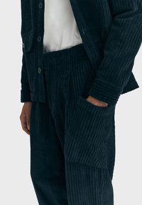Persona che indossa pantaloni in velluto a coste blu scuro e una giacca abbinata con una grande tasca, mano all'interno della tasca, camicia bianca sotto.